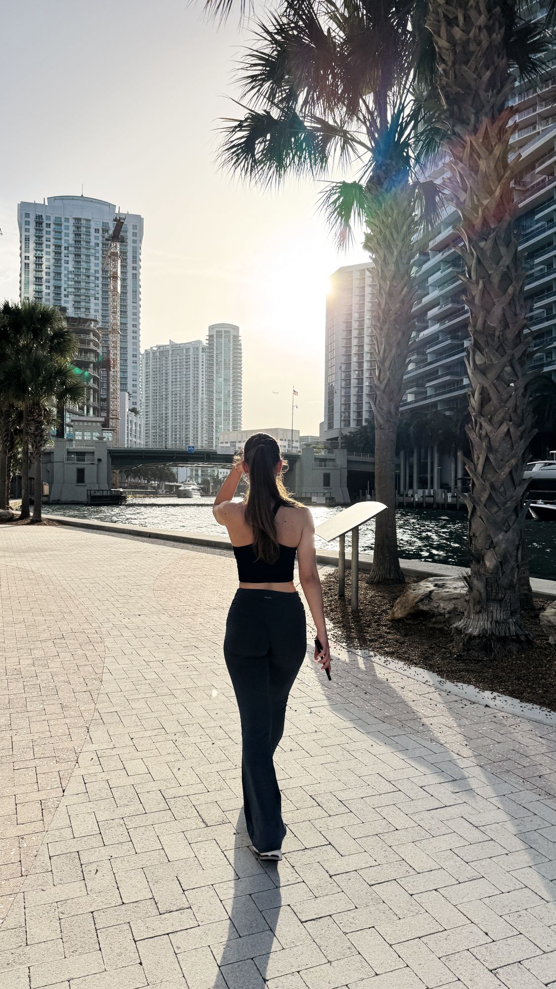 A figure walking a waterfront promenade lined with palms, backlit by the late afternoon sun in Dubai.